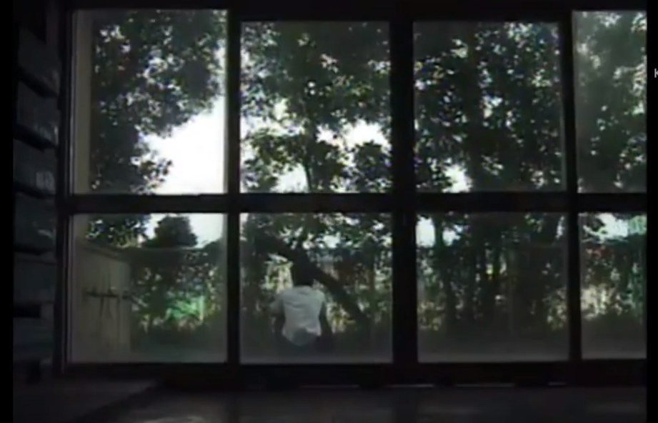 a shot of a young man's back taken from inside the building he sits on the steps of.