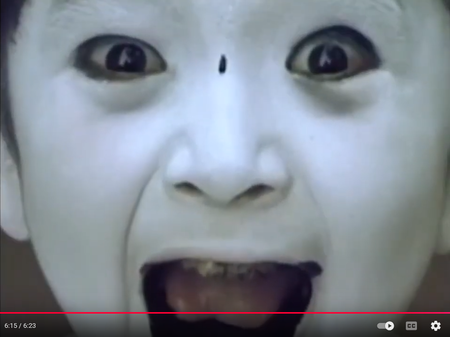 a young white-painted boy grinning at the camera as he spits black liquid from his mouth.